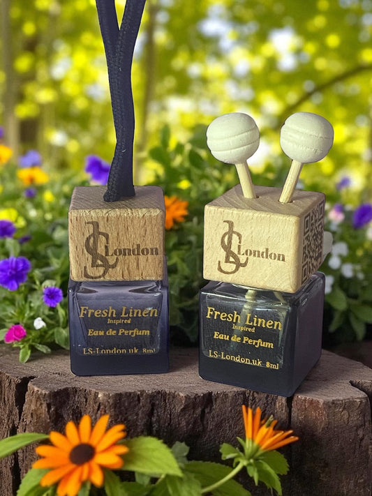 Two LS London Fresh Linen car diffusers Eau de Parfum bottles on a wooden block with flowers in the background. 